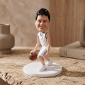 Basketball Player Bobblehead Figurine Custom basketball player bobblehead figurine dribbling a ball, wearing a white uniform with blue trim, displayed on a textured stone tabletop.