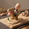 T-Rex Riding Child Bobblehead Figurine A bobblehead figurine of a child wearing a DKNY shirt and riding a brown T-rex on a sandy base, displayed on a stone slab.