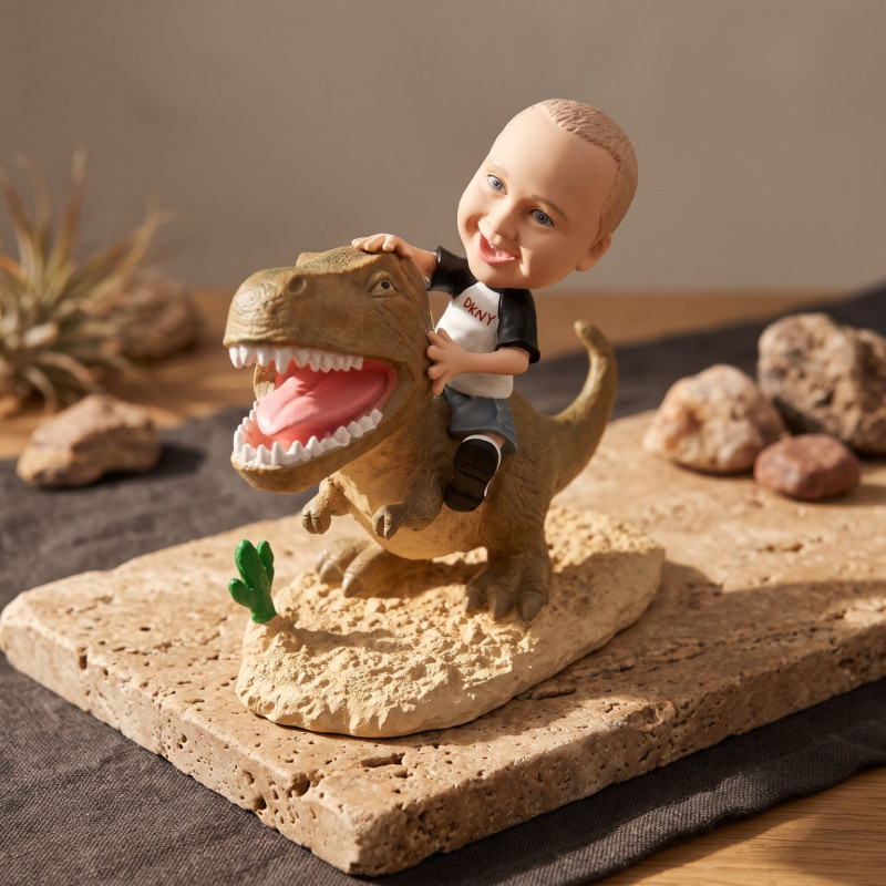 T-Rex Riding Child Bobblehead Figurine A bobblehead figurine of a child wearing a DKNY shirt and riding a brown T-rex on a sandy base, displayed on a stone slab.